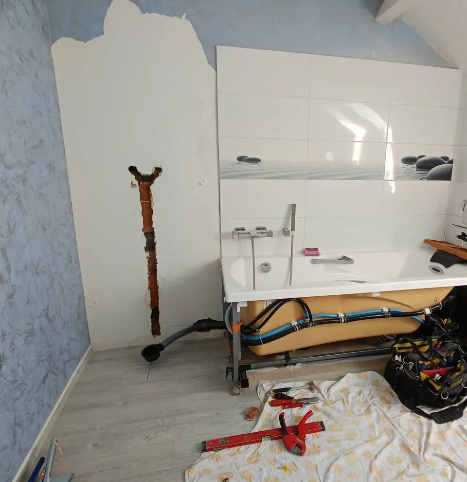 Chantier en cours : baignoire posée, alimentations en PER bleu visibles dessous, faïence blanche et outils posés sur une bâche