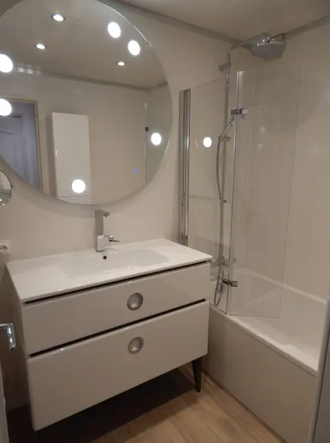 Meuble vasque blanc à deux tiroirs sous un grand miroir rond lumineux, baignoire avec pare-bain et colonne de douche
