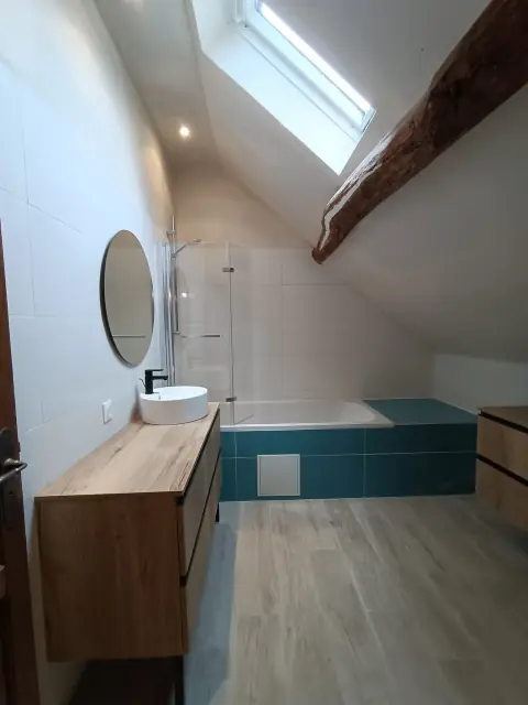 Salle de bains sous combles avec poutre apparente : baignoire habillée de carrelage bleu canard, vasque à poser sur meuble bois et miroir rond